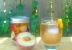 A canning jar of DIY peach liqueur next to a stirred cocktail and a fresh, halved peach.