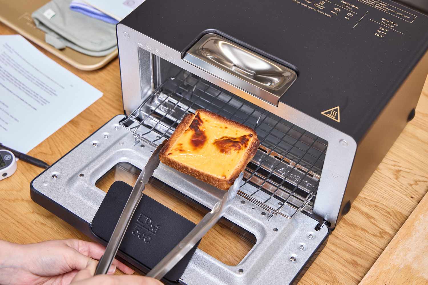 A balmuda toaster oven with a slice of toasted bread being removed using tongs