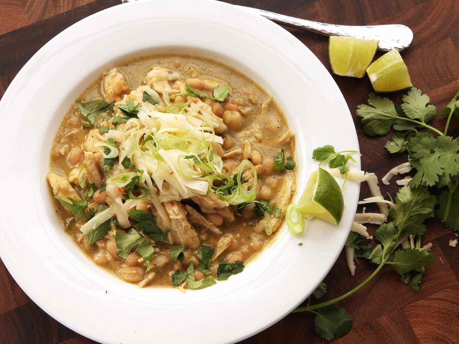 Overhead shot of white bowl of white chicken chili topped with shredded cheese, chopped cilantro and lime wedges.