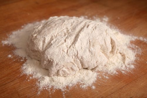 Soft bread dough covered in thick layer of flour on wooden cutting board