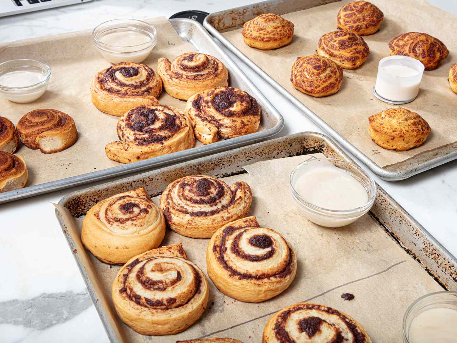 Cinnamon rolls arranged on baking trays with small cups of icing