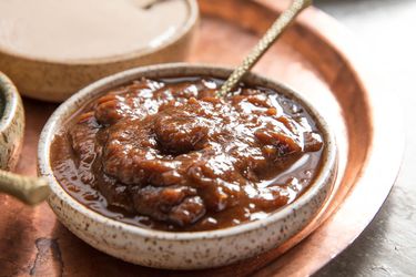 A wide, shallow cream-colored ceramic bowl with brown speckles of glaze on it, holding chunky tamarind chutney and a metal spoon. The bowl is placed on a copper tray and in the top left corner of the image is another similar bowl holding a different sauce.