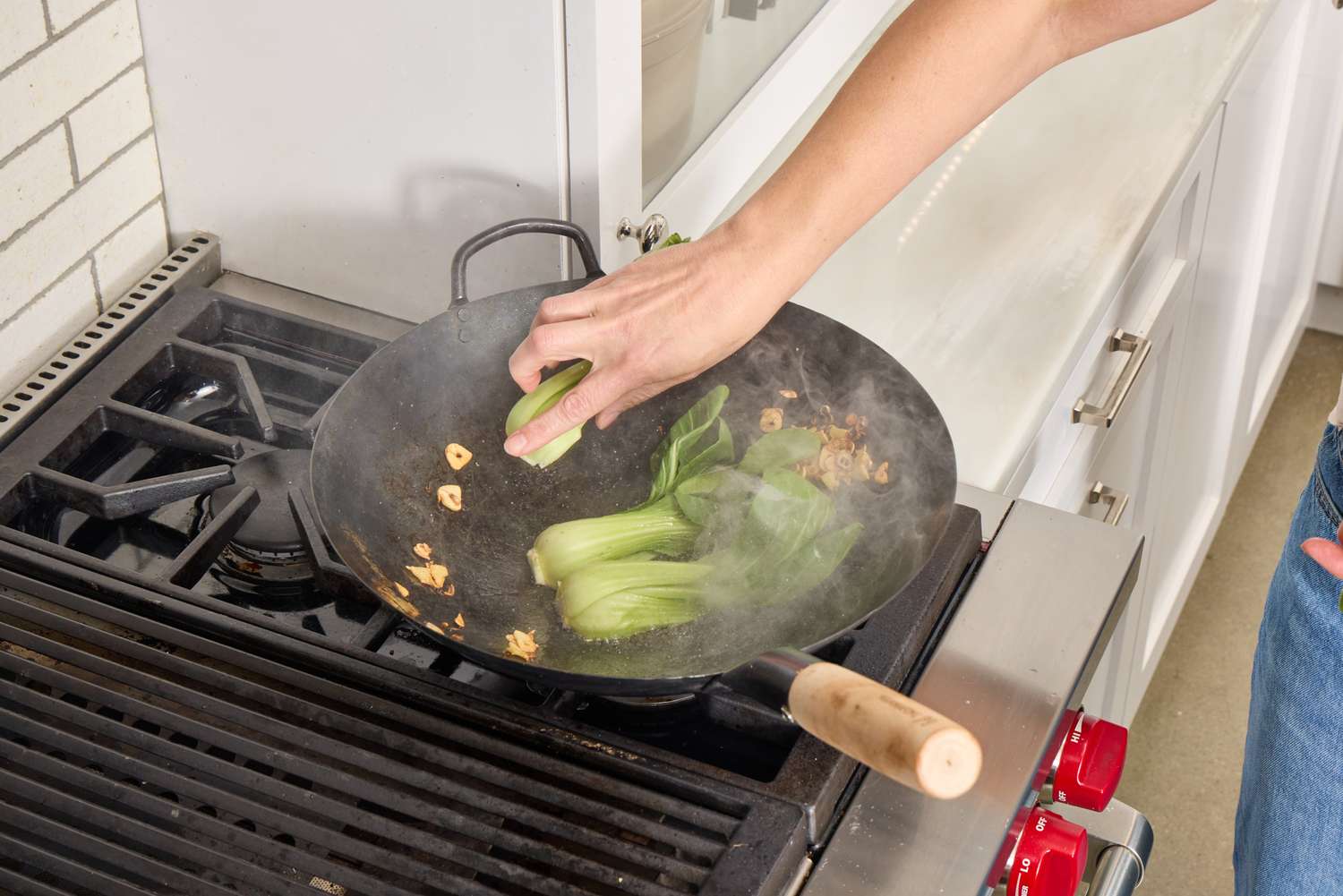 A person cooks bok choi in the Mammafong Pre-Seasoned Blue Carbon Steel Flat-Bottom Pow Wok