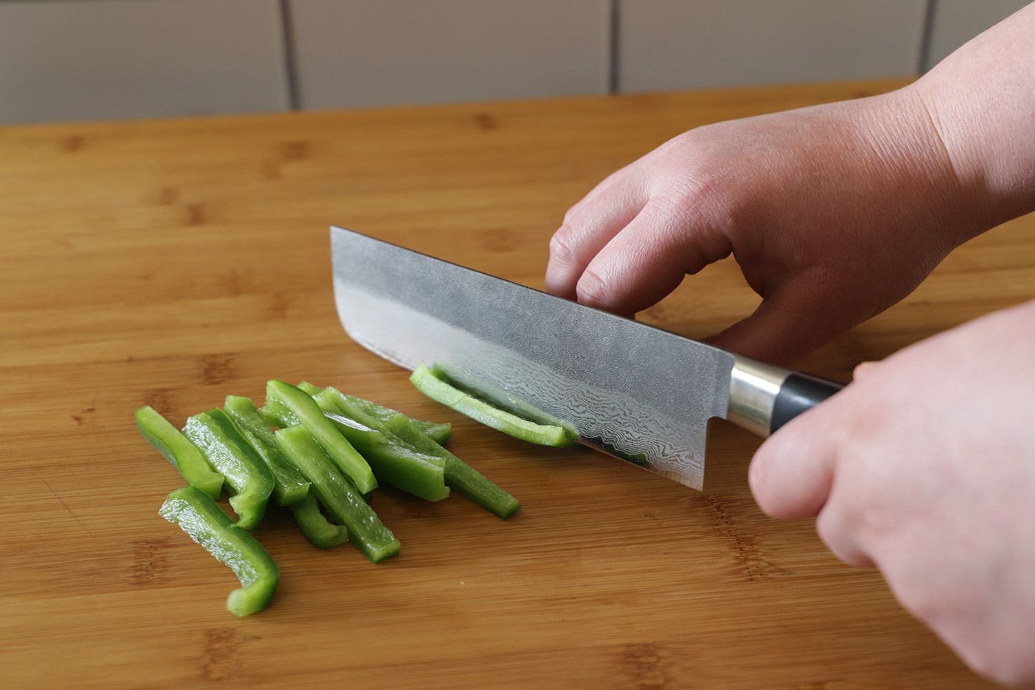 A person using a damascus steel nakiri knife to thinly slice green bell pepper on a wooden cutting board