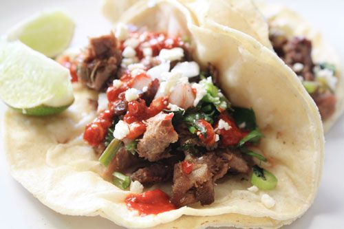 A fully-dressed lengua taco. The tongue is sprinkled with crumbled queso fresco, cilantro, and green onion. Salsa has been drizzled on top and lime wedges are close at hand.