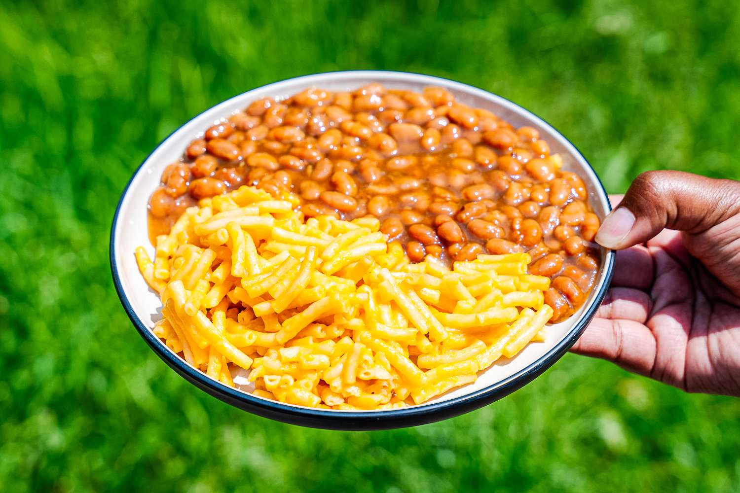 A hand holding an outdoor dinnerware plate full of baked beans and macaroni and cheese