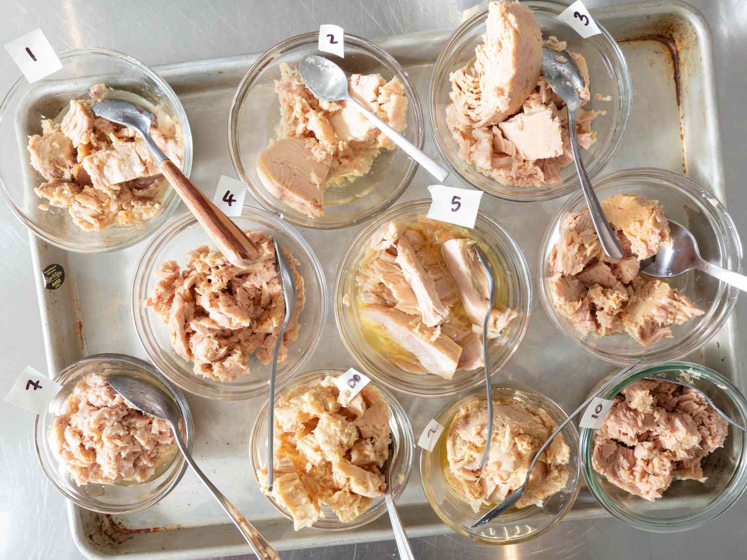 Many types of tuna in oil in small bowls side by side on a sheet pan from above 