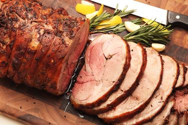 A slow-roasted leg of lamb sliced on a cutting board, flanked by rosemary sprigs, peeled garlic cloves, and wedges of lemon.