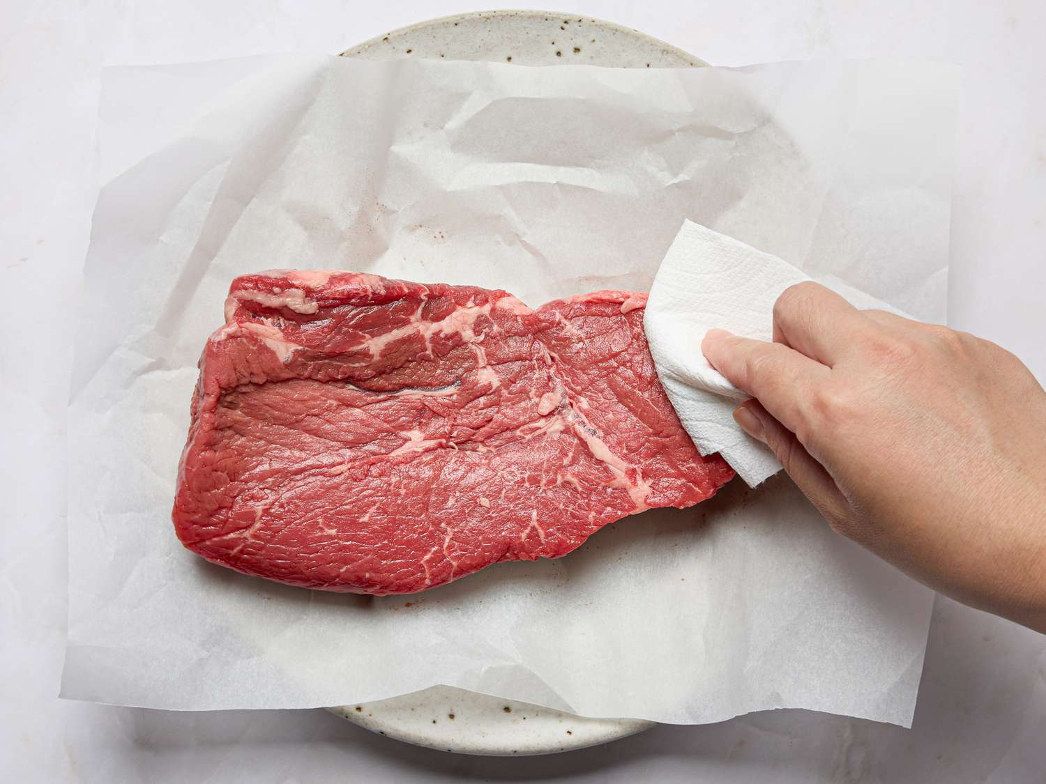 A piece of raw steak being patted dry with a paper towel on parchment paper