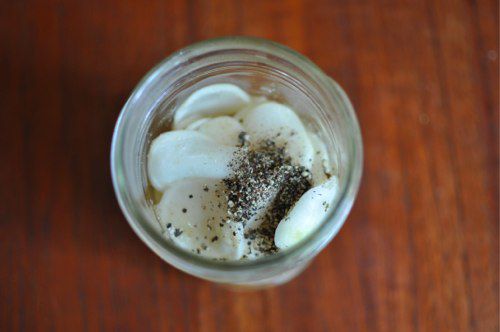 Overhead view of a pint jar of the salted turnips, covered with a pinch of ground black pepper, ready to be filled with brine.
