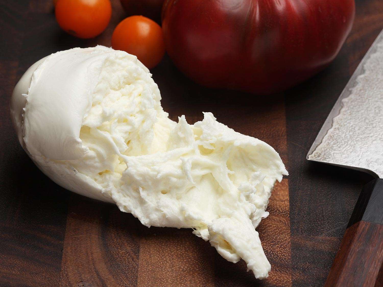 A torn mozzarella ball on a wooden work surface, next to tomatoes mostly out of frame. 