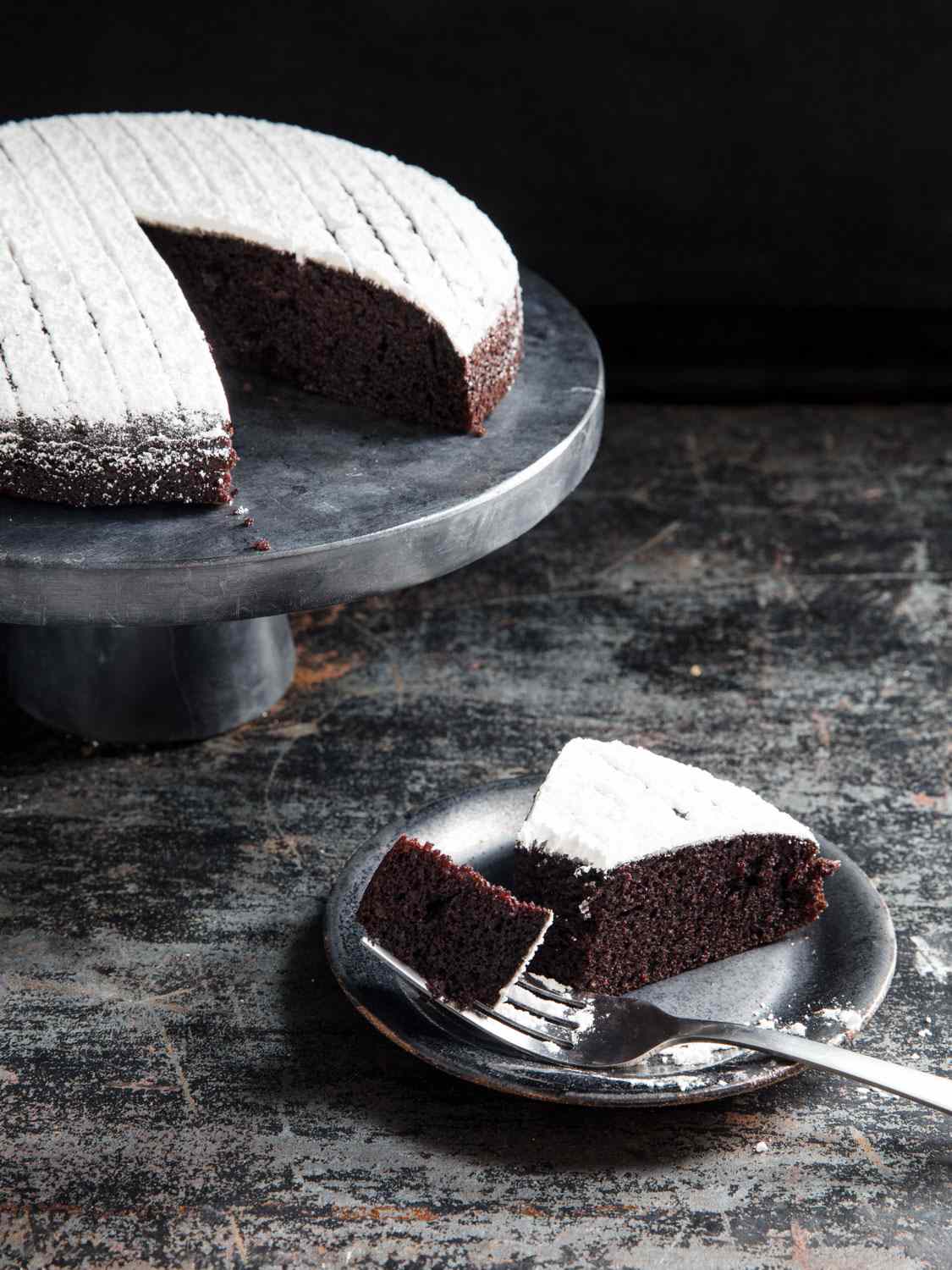 A slice of chocolate cake with the cake in background on a stand.
