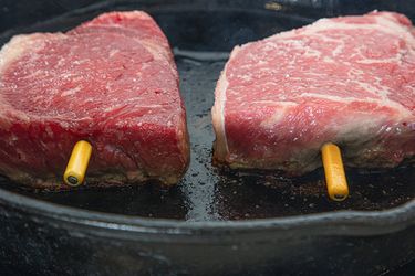 Two pieces of steak being cooked in a castiron skillet with yellow thermometers inserted
