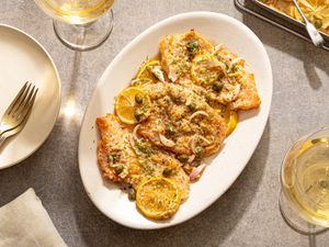 A plate of chicken piccata with lemon slices and capers on a table next to glasses of white wine
