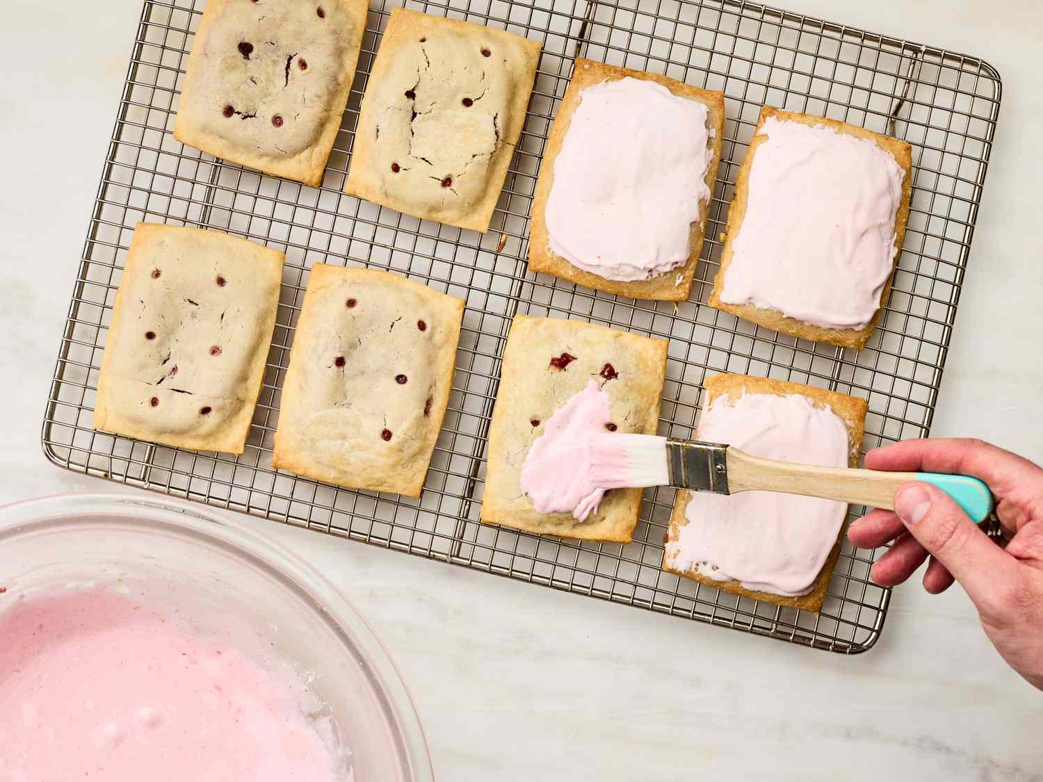 A hand icing pastries on a cooling rack filled with strawberry rhubarb Pop-Tarts
