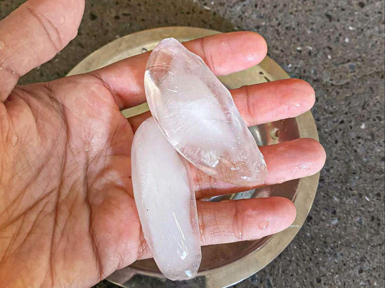 Two clear ice pieces in a hand above a metal plate