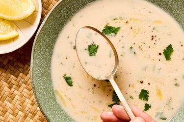 A bowl of garlic soup with garnish and a hand holding a spoon over the surface a plate of lemon slices to the side