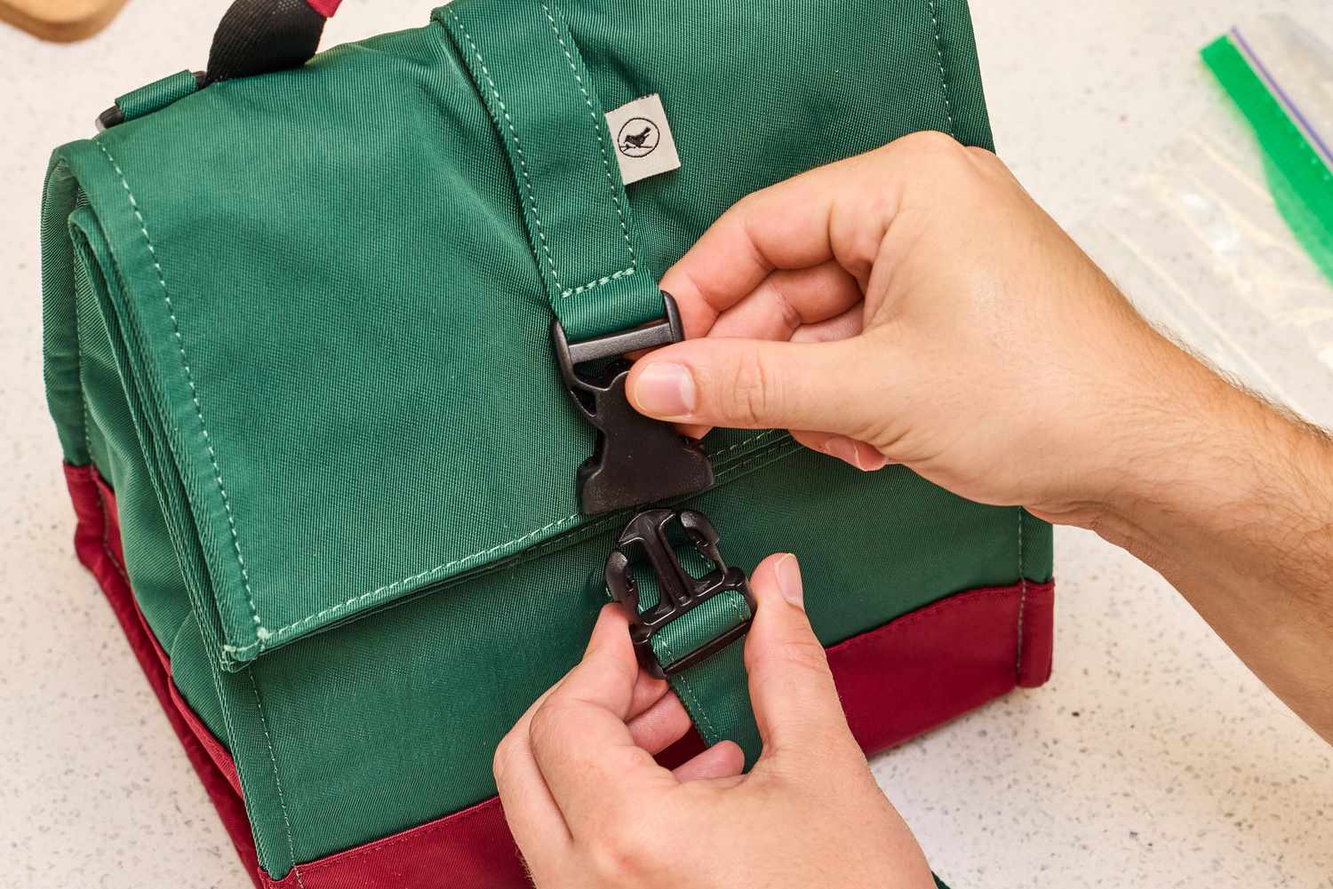 A person closing a buckle latch on a green and red roll-top lunch bag
