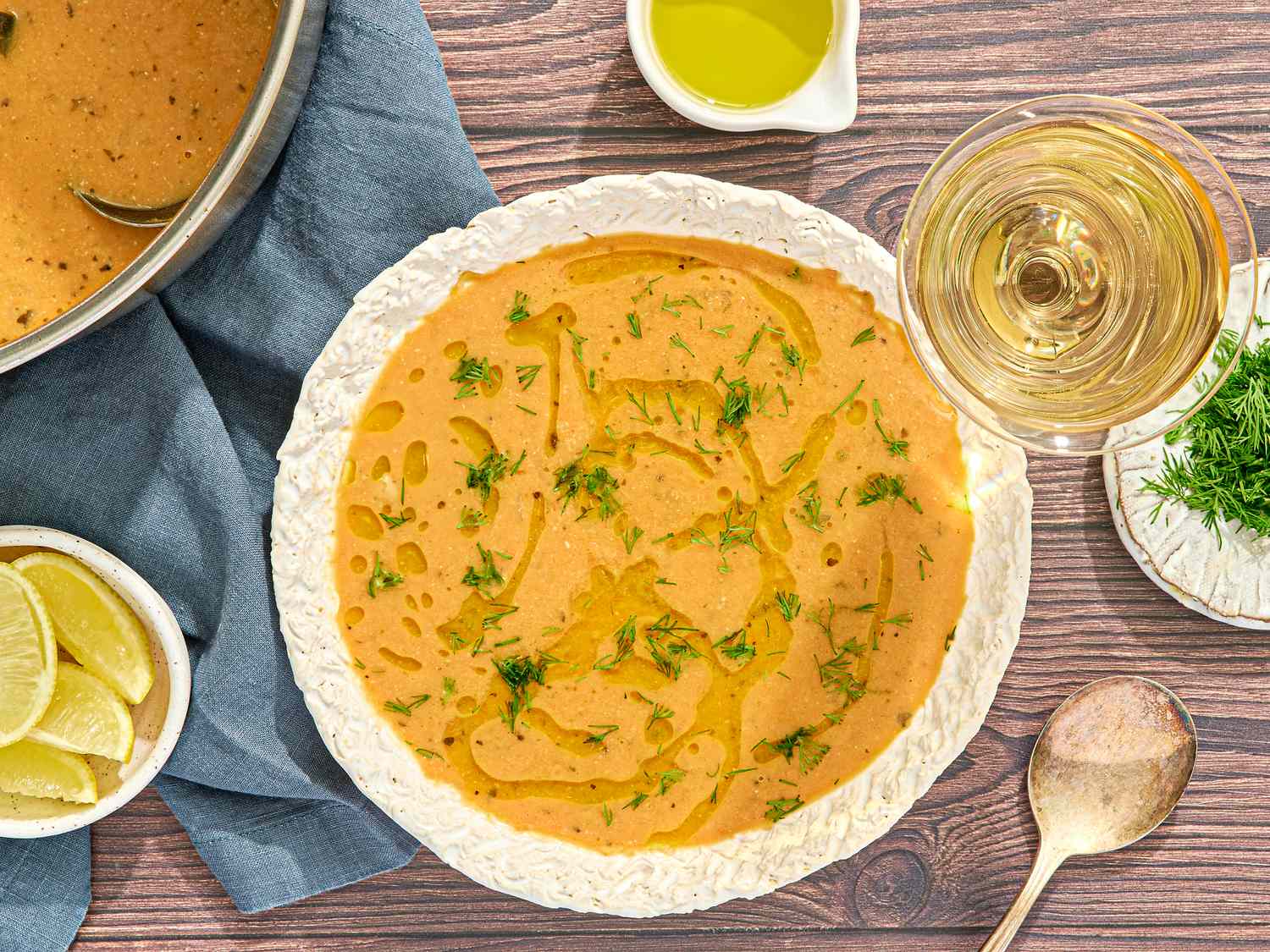 A bowl of chickpea stew garnished with herbs accompanied by a glass of wine lemon wedges and a wooden spoon