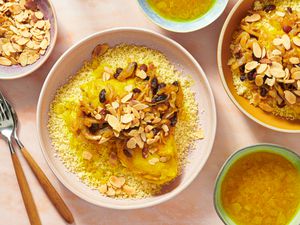 2 plates of Chicken couscous T'faya on pink surface surrounded by bowls of broth, toasted almonds, and forks