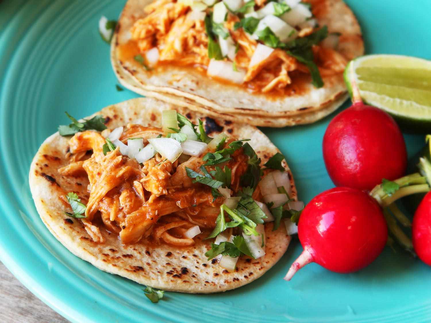 Two tacos filled with chicken tinga on a plate with some radishes and lime wedges. 