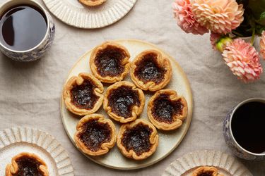 Overhead of platter with Canadian butter tarts. 3 smaller plates have one tart each. Coffee and flowers on the tabletop, with linen tablecloth.