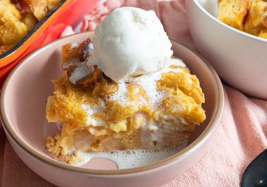 Soft pink bowl with bread pudding a la mode, on a soft pink textile, with a white bowl of bread pudding and full red tray of bread pudding on the sides. 