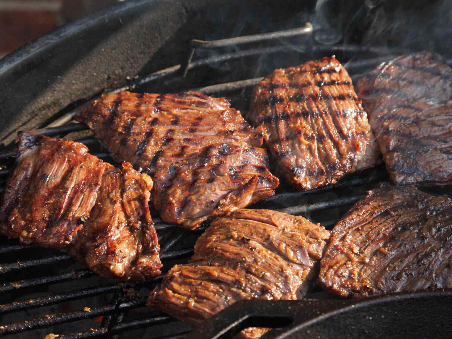 Skirt steaks for fajitas, in various stages of doneness, cooking on a grill next to a cast iron skillet.