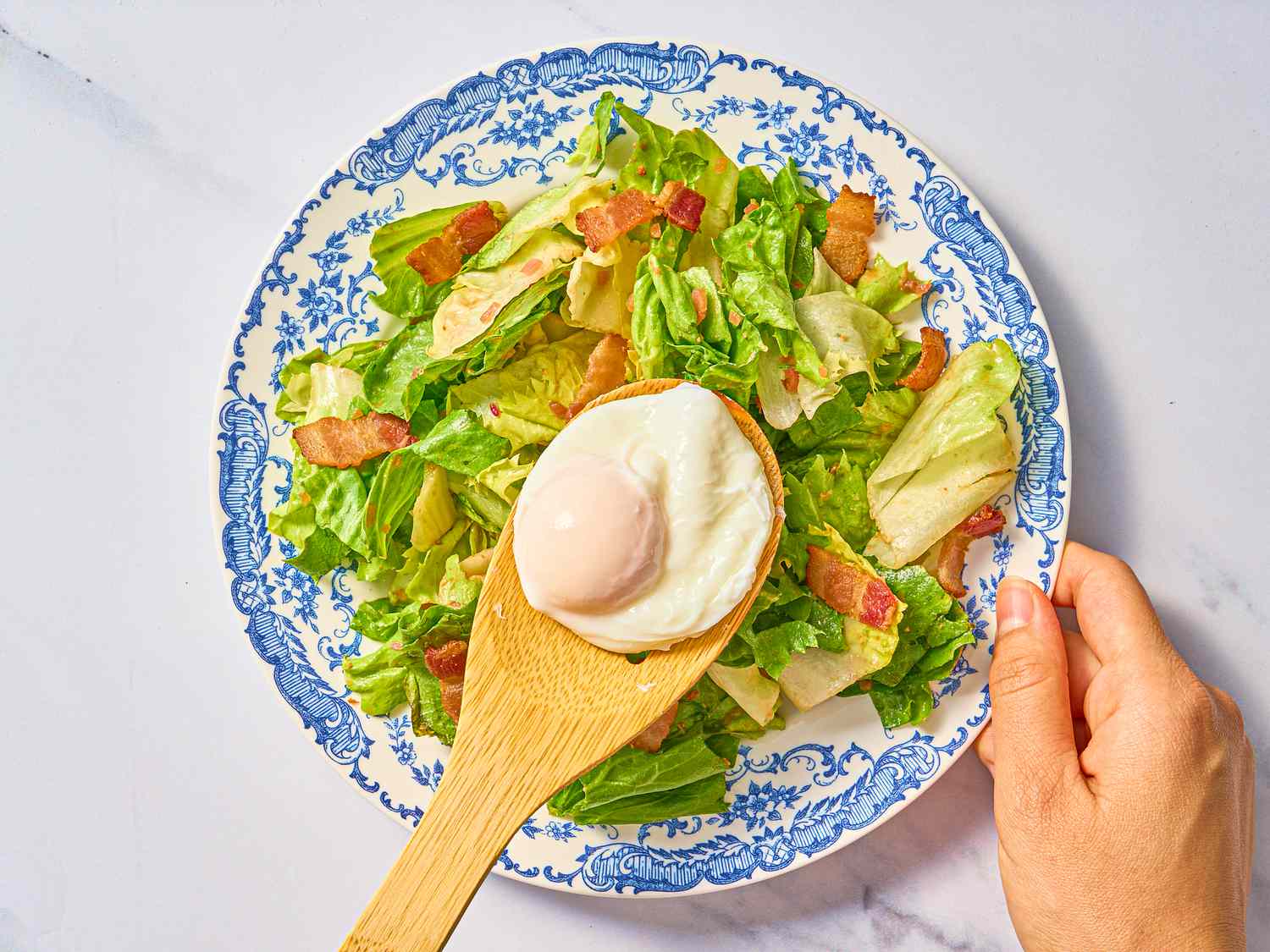 Poached egg placed on top of Salade Lyonnaise an old fashioned blue and white plate, on a white surface 