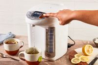 A hand pressing a button on an electric water boiler while hot water fills a mug surrounded by tea and sliced lemon