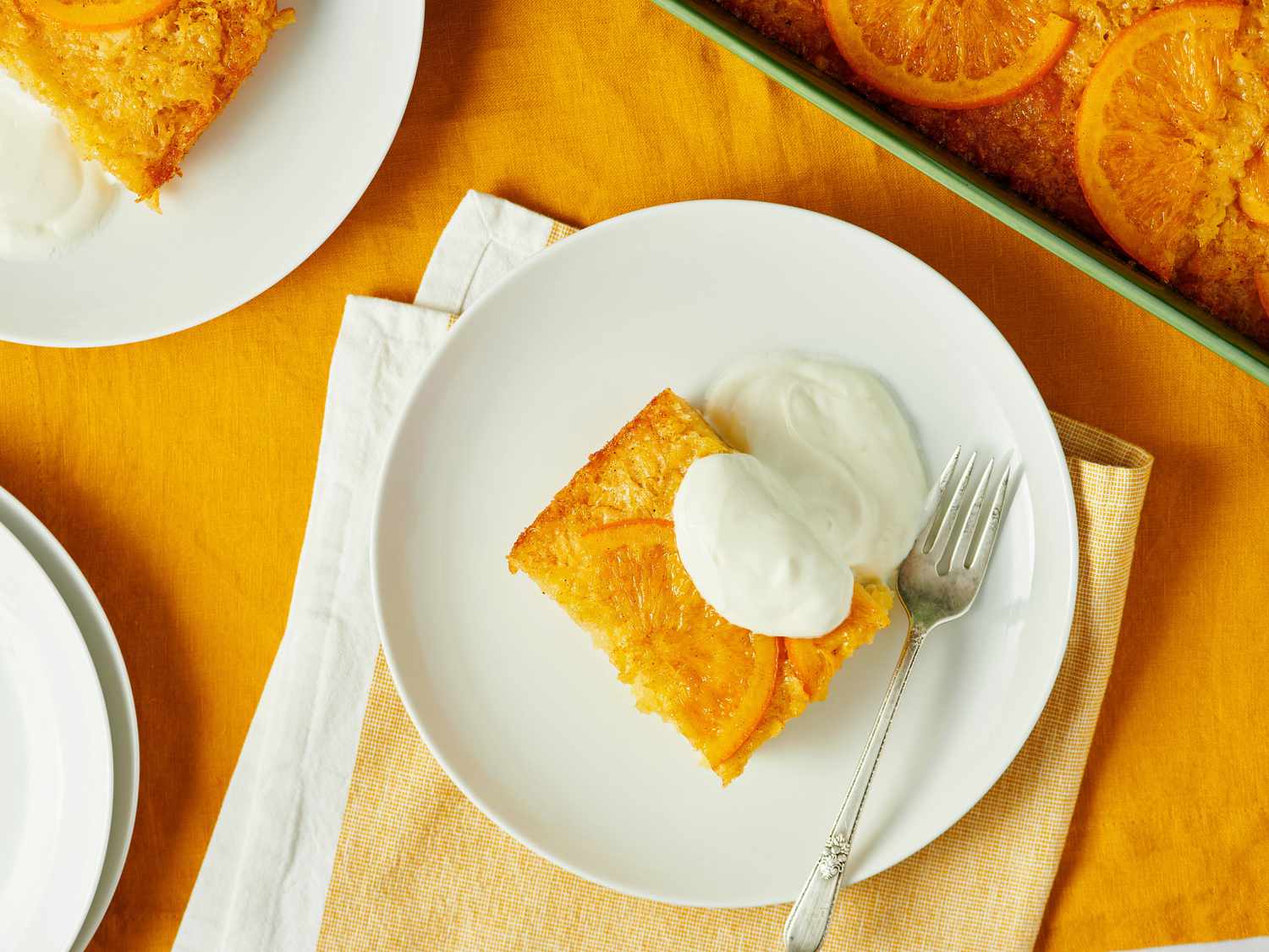 A plate with a slice of orange-infused portokalopita garnished with a dollop of cream, served with a fork