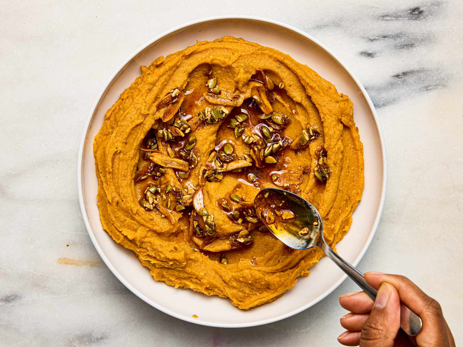 Sweet potato pepita dip garnished with toasted seeds and a drizzle of sauce a hand holding a spoon near the dish