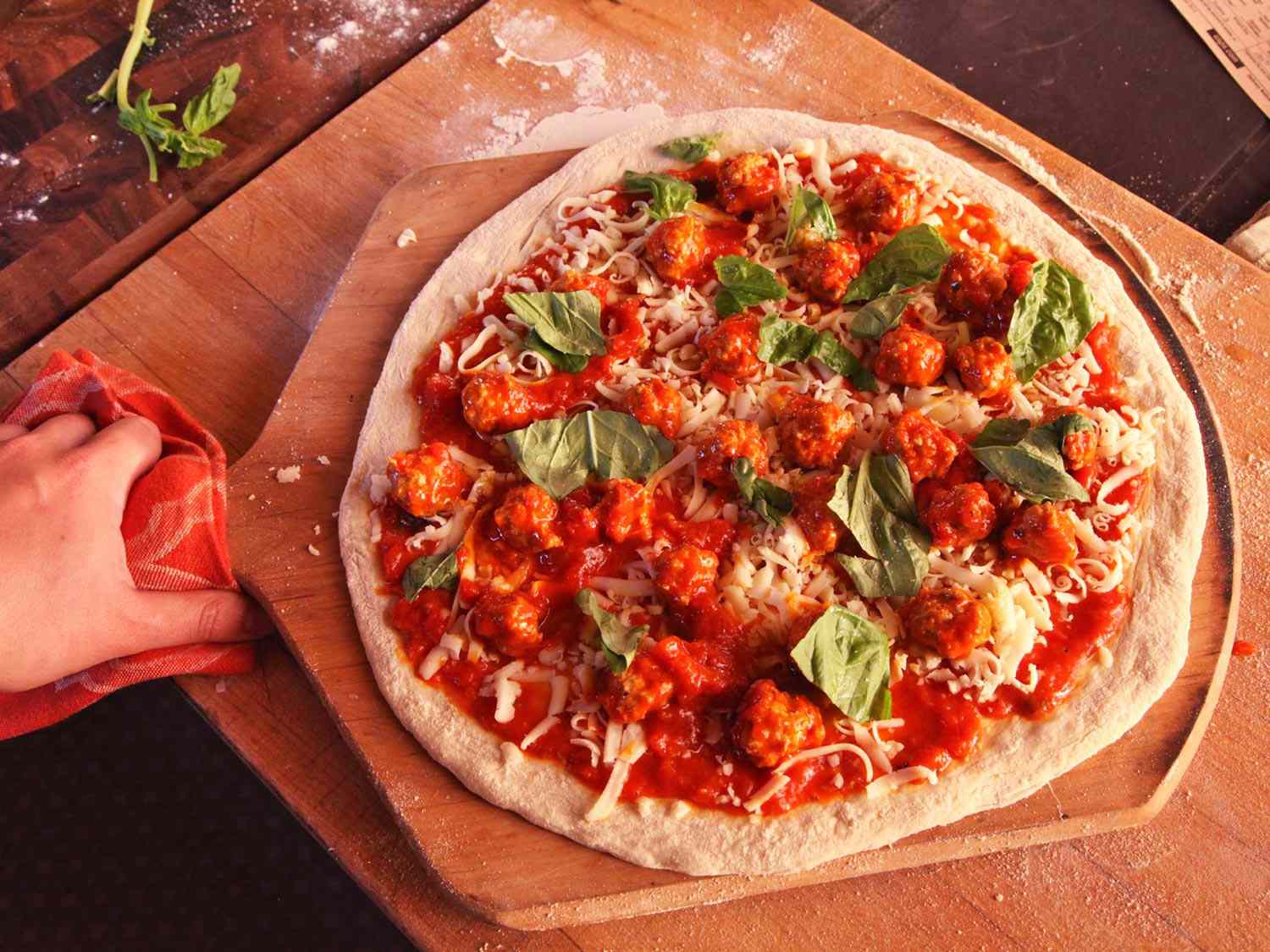 A fully topped meatball pizza with scattered basil leaves, on a wooden pizza peel.