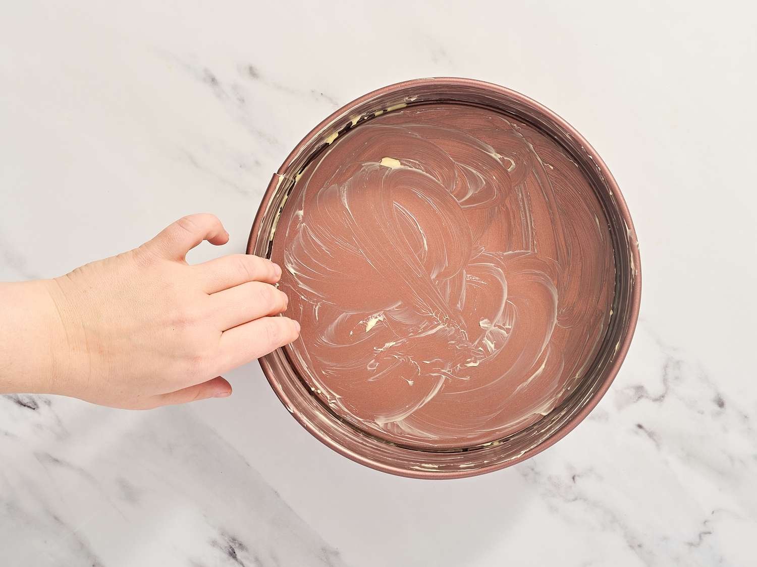 A hand greasing a round cake pan with butter or a similar substance preparing it for baking