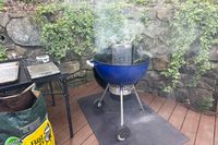 A charcoal grill with a chimney starter emitting smoke situated on a wooden deck near ivycovered stone walls with grilling supplies nearby