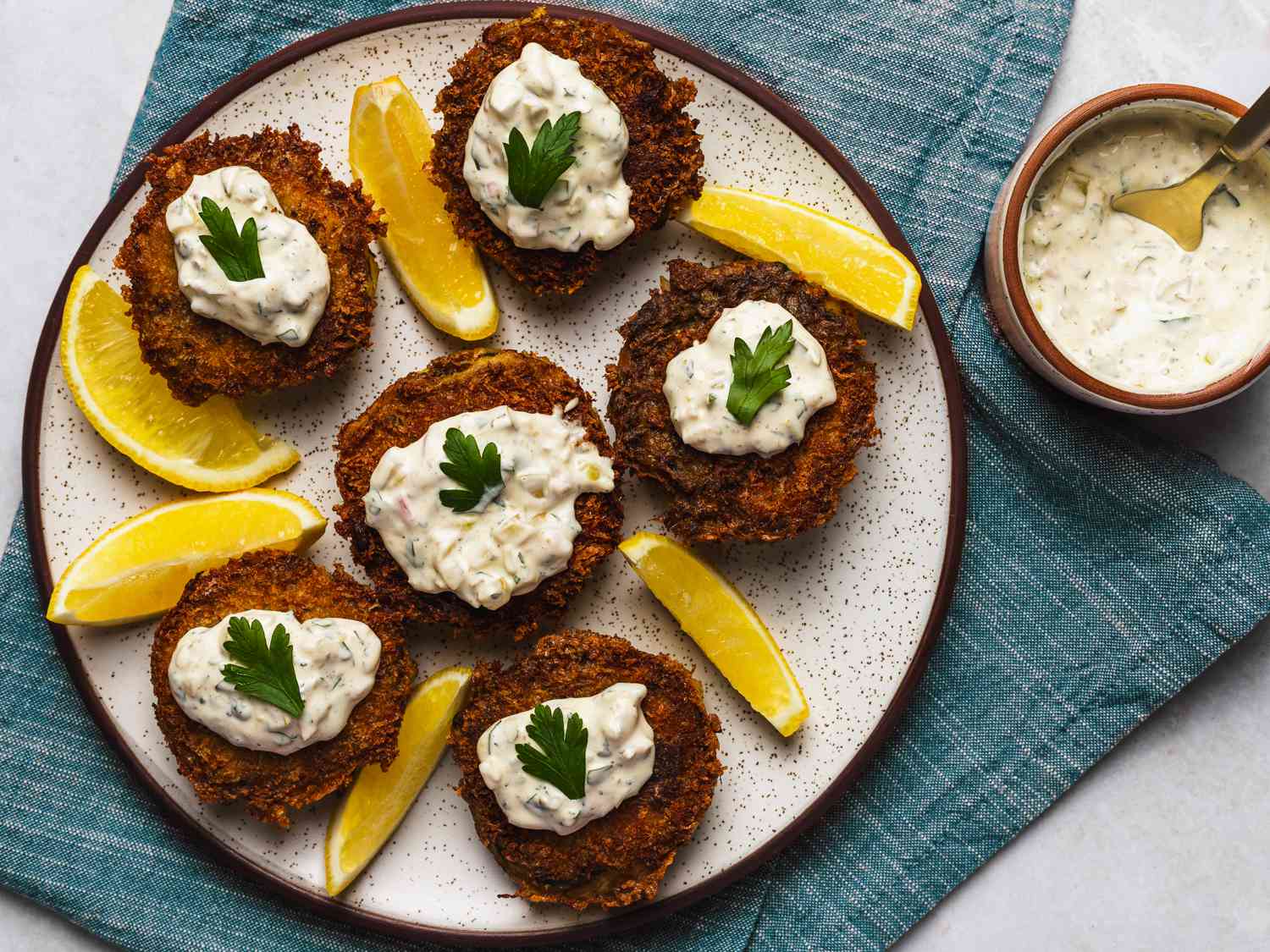 Six crispy herbed crab cakes, each topped with tartar sauce and a single leave of flat parsley, interspersed with slices of lemon. The plate is on a blue textured cloth, and there's a small bowl of tartar sauce and a spoon in the top right corner of the image.