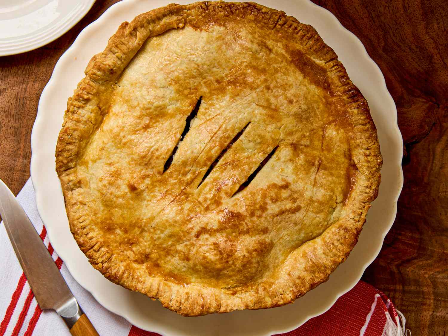 A baked pie with slitted top crust on a white plate set on a wooden table with a redstriped napkin and a knife nearby