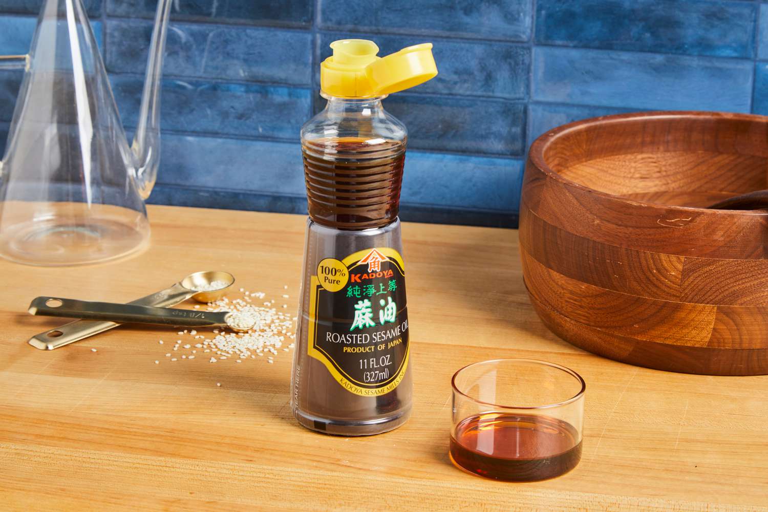 Kadoya roasted sesame oil bottle on a wooden table, next to measuring spoons and a glass container of oil