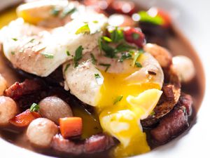 Close-up of yolk running out of poached egg in pool of red wine sauce with carrots, pearl onions, and bacon
