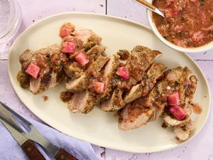 sliced pork with rhubarb sauce drizzled on it, on aa cream colored plate, with bowl of rhubarb sauce to the side, on a tile surface.
