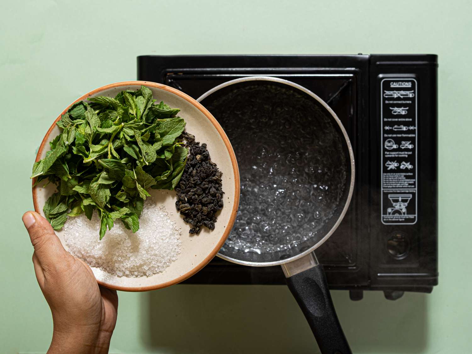 A hand holding a plate with mint leaves, sugar, and tea leaves over a boiling pot on a stovetop