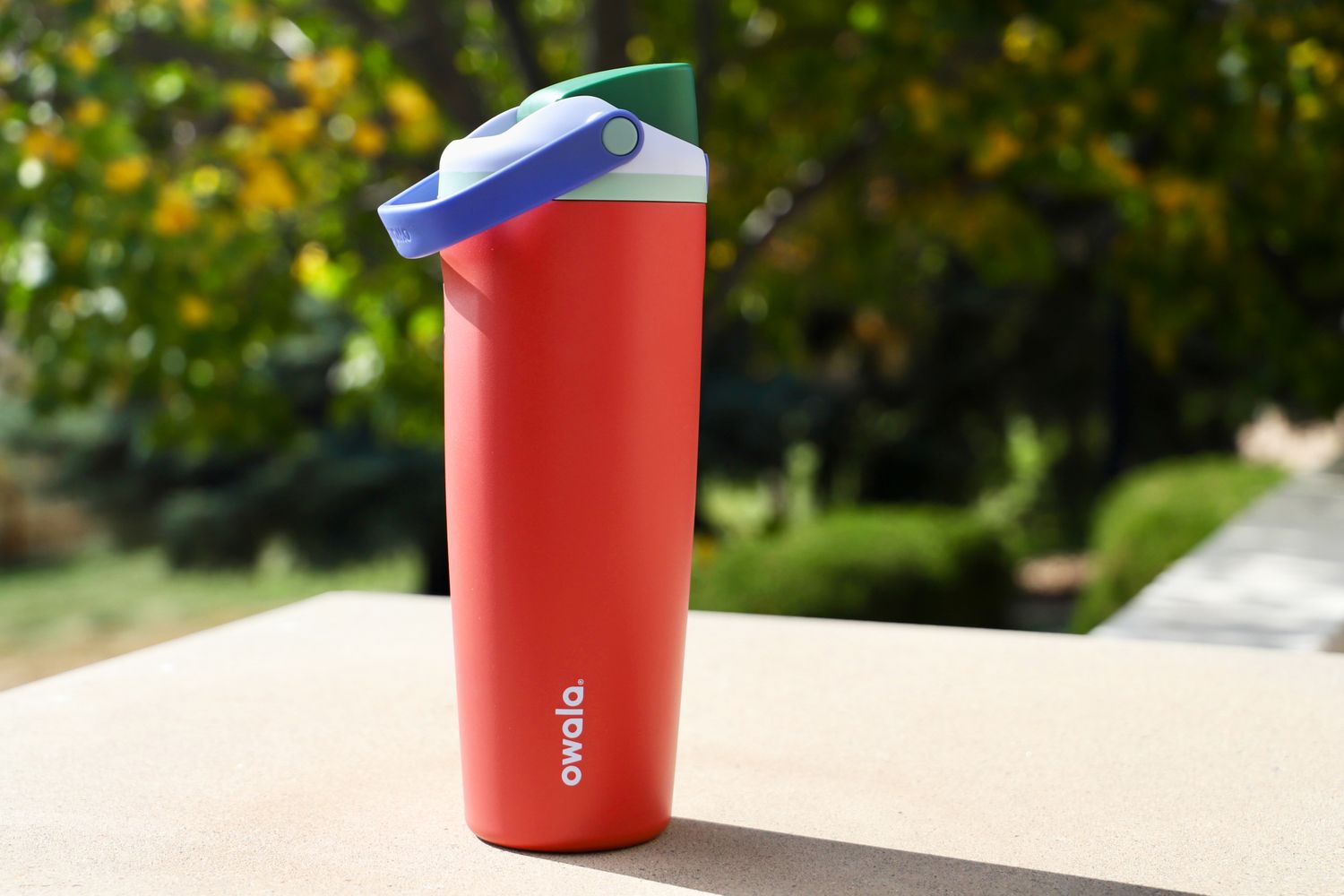 An Owala FreeSip Sway (in Summer Harvest colors) photographed outside against a lush tree background.