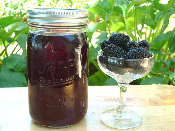A quart mason jar of homemade blackberry liqueur next to a coupe glass filled with fresh blackberries.
