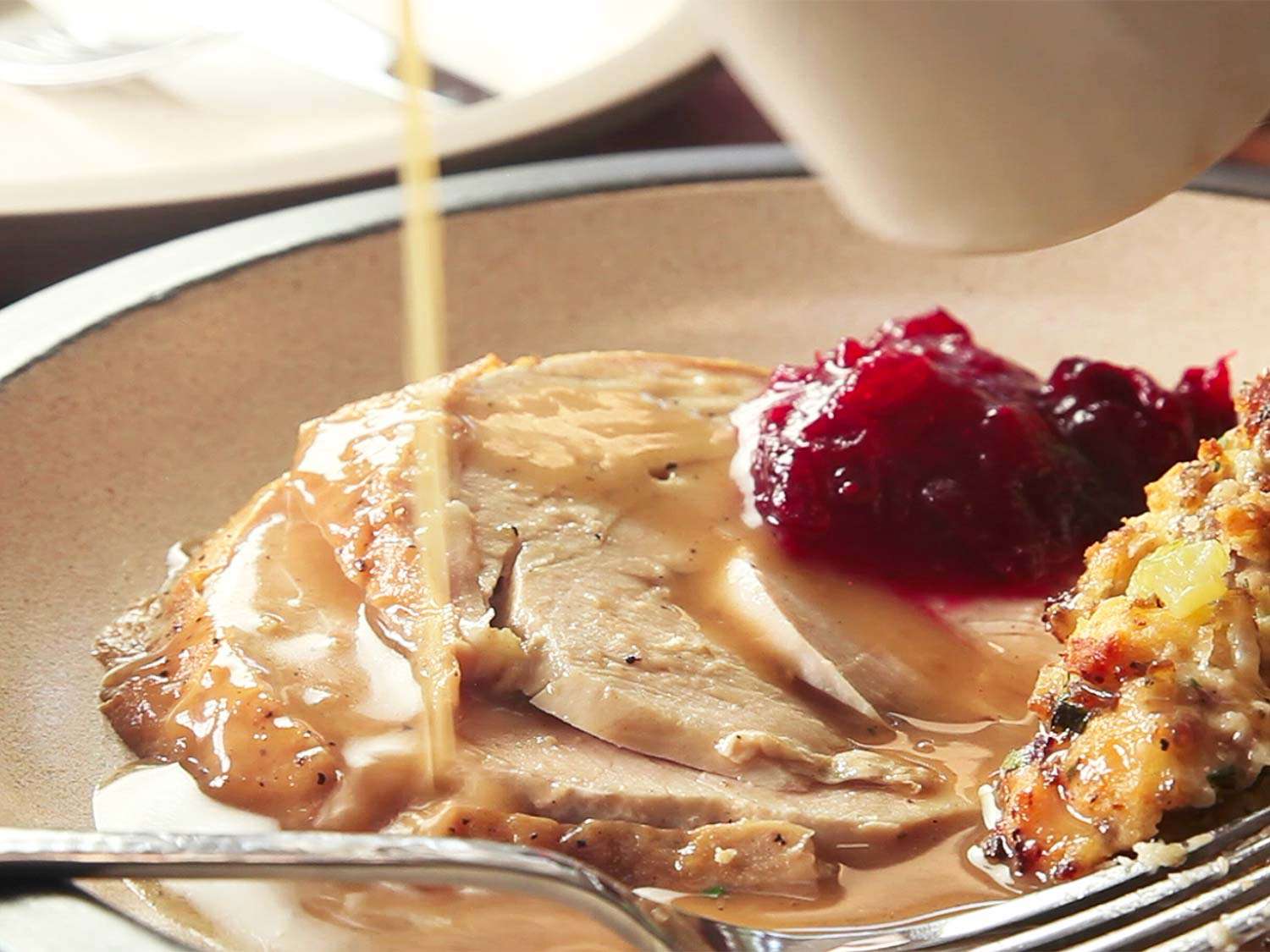 Pouring gravy from a pitcher over a plate of sliced roast turkey meat, cranberry sauce, and dressing.