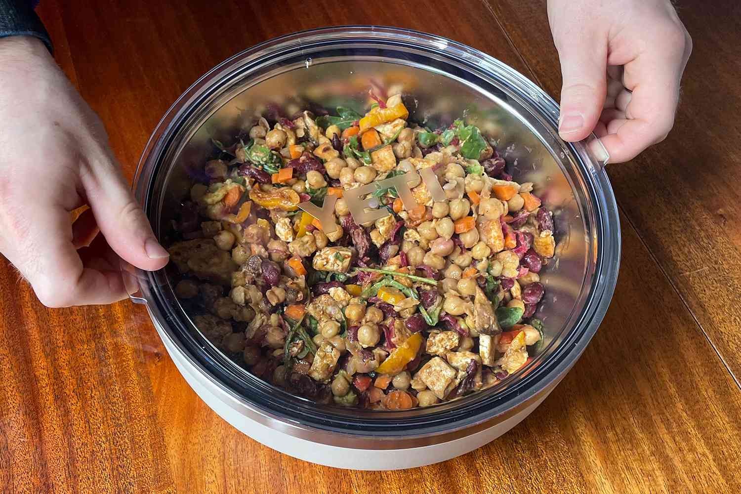 Hands holding a bowl of mixed salad with chickpeas and vegetables on a wooden surface