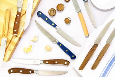 a variety of steak knives on a white tile surface with garlic cloves and mushrooms
