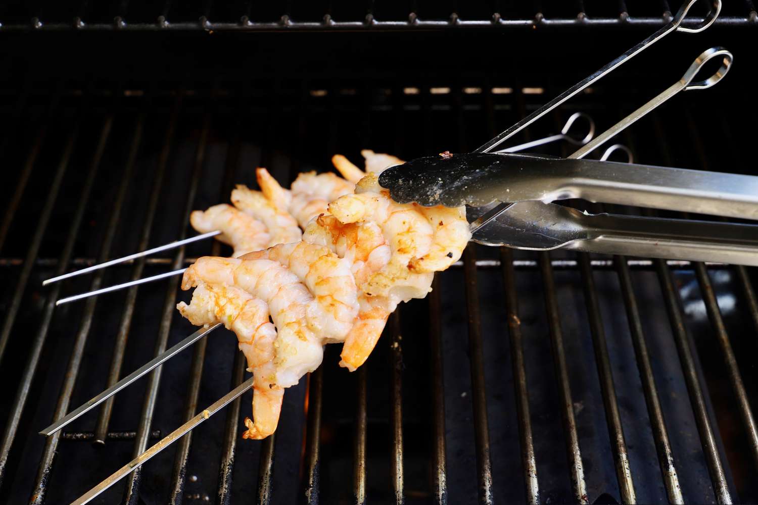 A closeup look at tongs flipping two metal skewers that have shrimp threaded onto them