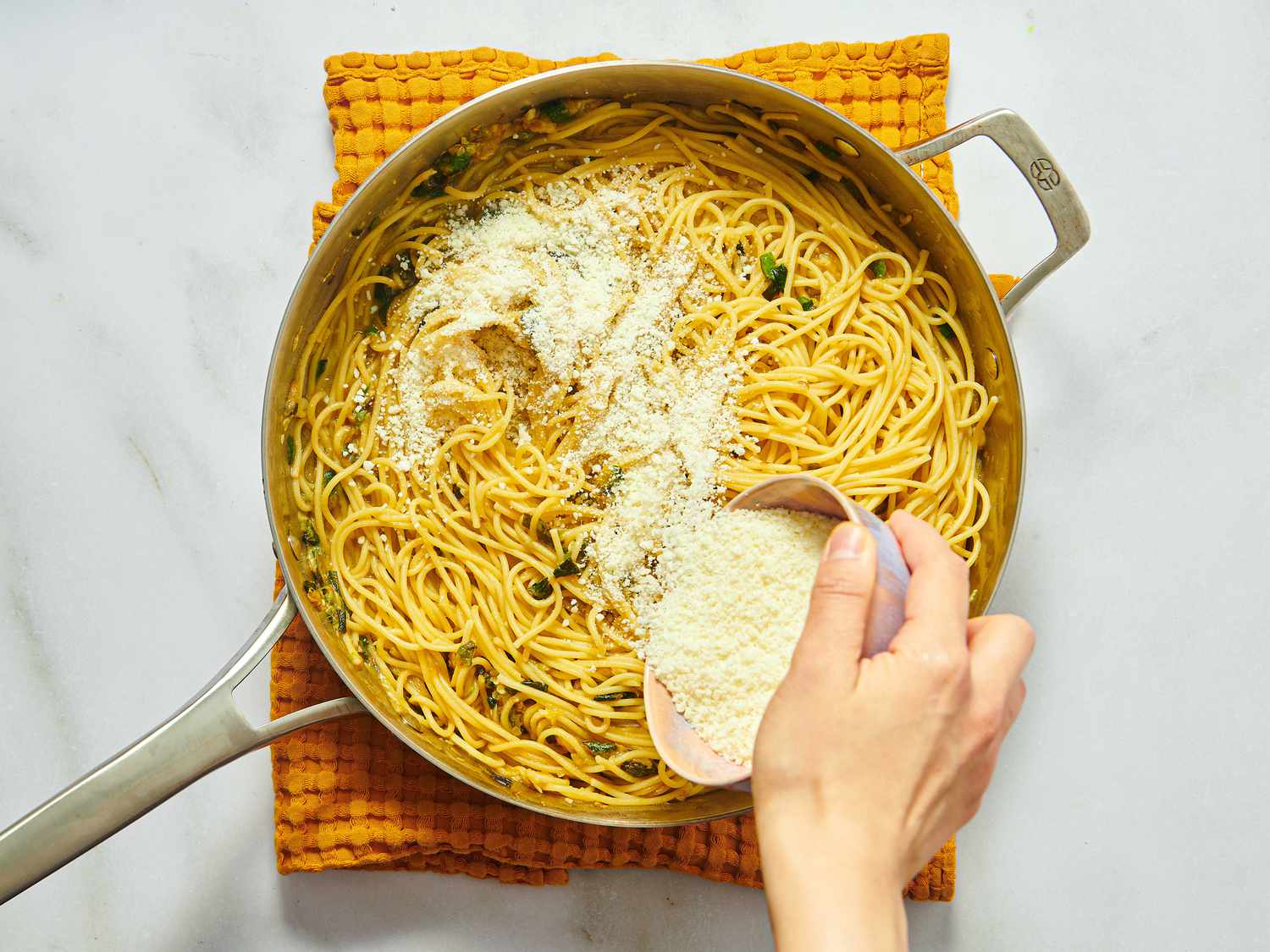 Hand sprinkling grated cheese over spaghetti in a skillet on a textured surface