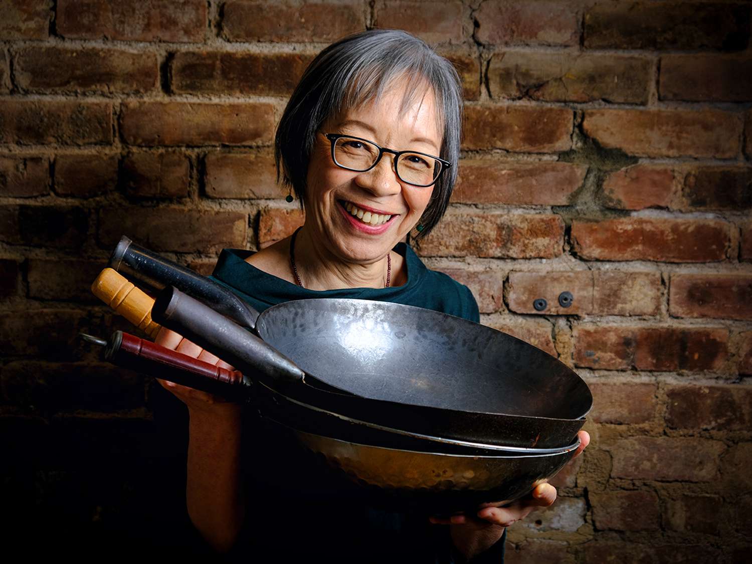 Portrait of Grace Young holding a stack of woks against a brick wall