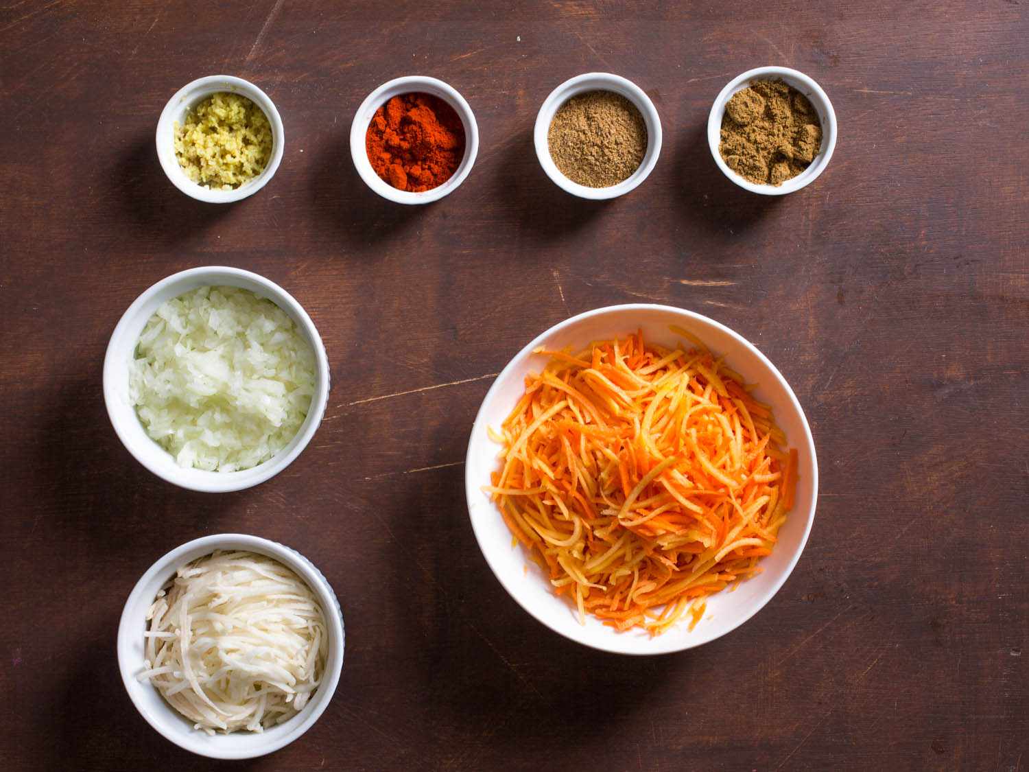 Overhead shot of ingredients for sweet potato, squash, and carrot latkes: shredded carrot, squash, and sweet potato; shredded potato; chopped onion; fresh ginger; paprika; cumin; and coriander